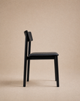 Here’s a revised version of your sentence using the provided product data: The Poise Dining Chair | Leather by Wooden Stories, featuring a modern black wooden frame with a cushioned leather seat and angled backrest, is shown in profile against a plain beige background.
