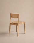 A minimalist solid oak Poise Dining Chair from Wooden Stories’ Danish Collection, featuring a curved backrest and no armrests, positioned at an angle on a plain beige background.