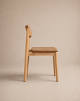 The Poise Dining Chair | Leather by Wooden Stories, featuring a solid oak frame and a tan leather cushioned seat with a simple, modern design, is shown in profile against a neutral beige background.