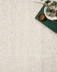 A hand-woven white textured Sloane Rug - Sky by Loloi is shown with a green book, a white plate with leaves, and a small brown dish arranged in the top right corner.