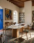 Sunlit study with a Tathra Ellipse Dining Table | Stone Top by Wooden Stories, wooden chairs, book-filled shelves, colorful artwork, patterned tile floor, and a bright blue door. Rolled papers and books are scattered on the table.