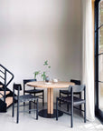 A SOBU Posta Dining Table in white oak with a satin black steel base is paired with four black chairs beside sheer-curtained windows. A small plant, cup, and bowl sit on the table, while black metal stairs accent this minimal modern space.