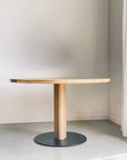 The Posta Dining Table - White Oak by SOBU features a minimalist round solid white oak top with a cylindrical base and satin black steel footing, shown in a bright room with light walls and floors.