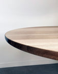 A close-up of the rounded edge on the SOBU Posta Dining Table - White Oak, featuring a satin black steel base, set against a white wall and dark carpeted floor.
