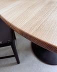 Close-up of the SOBU Posta Dining Table - White Oak, highlighting its visible grain detail beside a dark chair and round satin black steel base on a light floor.