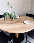 The SOBU Posta Dining Table - White Oak features a solid oak top and satin black steel base. On the round surface are a potted plant, beige bowl, and white plate, with two black chairs nearby against a light wall.