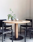 A SOBU Posta Dining Table in white oak with a satin black steel base is paired with four black chairs. On the table are a potted plant, a white bowl, and a plate. The minimalist setting features neutral-colored walls.