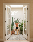 A bright room with French doors opens to potted plants—a tall cactus and aloe on a wooden stand—beside the SOBU Blix Counter Stool, all arranged on a pebble-patterned floor beneath a skylight.
