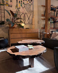 Certainly! Here’s a rewritten version of your sentence using the product data: A Bolinas Nesting Coffee Table Set in Claro Walnut by SOBU, handmade in Oakland with natural edges, holds a stack of magazines and small dishes. Sunlight filters through, highlighting the rich wood grain. Modern furniture and a patterned wall hanging appear in the background.