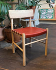 A SOBU Cesco Dining Chair in solid white ash with a heritage red leather seat sits on a dark wooden floor, surrounded by potted plants and colorful framed artwork in the background.