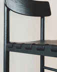 Close-up of the SOBU Cesco Dining Chair - Black Ash, featuring a curved backrest and a seat with woven leather straps, set against a plain white wall.