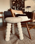 The Chubby Stool - Ash by Robert Saltonstall, with rounded textured legs and a Scandinavian soap finish, sits beside a woven lounge chair topped with a black and white pillow in a cozy modern living room accented by earthy tones and abstract art.