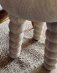 Close-up of rounded, spiral-carved wooden legs from the Robert Saltonstall Chubby Stool - Ash, resting on a textured beige woven rug.
