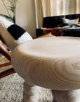 Close-up of the Robert Saltonstall Chubby Stool - Ash, featuring a smooth ash finish and rounded edges; in the background, a textured rug, modern chair with black-and-white cushion, and a sofa with throw pillows in warm natural light.