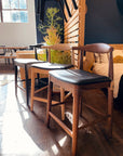 Three SOBU Copenhagen Counter Stools in Black Ash are lined up on a wooden floor in a sunlit room, with sunlight streaming through a large window. A tall plant and a bed can be seen in the background.