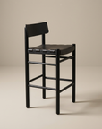 The Cesco Counter Stool by SOBU features a modern black finish in solid American ash, paired with a flat top grain leather seat, a low rectangular backrest, and simple straight legs. It is set against a plain beige background.
