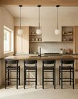 A modern kitchen with light wood cabinets, a large island with four SOBU Cesco Counter Stools in Black Ash featuring top grain leather seats, pendant lights above, open shelves filled with dishes, and a window letting in natural light.