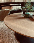 A close-up of the SOBU Disco Round Coffee Table in White Oak with Osmo oil finish holds a glass of water, green candle, small black decorative figure, and a book; a textured rug and cushioned sofa are in the background.