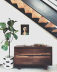 The SOBU Jupiter Sideboard - Walnut 2-Door sits under a staircase, topped with a white bowl and decor. A potted fiddle leaf fig and a portrait painting on the wall complete this minimalist, stylish interior scene.