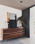 A modern entryway features the SOBU Jupiter Sideboard - Walnut 3-door, an abstract painting, glass sculpture, potted plant, striped wooden ceiling, gray rug, and white walls for a clean, contemporary look.