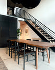 A modern dining area features the SOBU Mars Live Edge Dining Table in walnut with wood legs, black chairs, black staircase railings and cabinets, a plant, and framed art on a white wall filled with natural light.