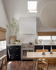 A modern kitchen features dark cabinets, light wood chairs, a SOBU Mars Live Edge Dining Table in white ash, a paper lantern pendant, skylight, potted greenery, and windows with woven shades that brighten the clean, airy space.