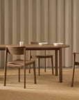A minimalist dining room with a Tolv wooden table, three Wooden Stories Poise Dining Chairs in Espresso Oak, white bowls and cups on the table, beige carpeting and vertical beige paneling. A partially open white door appears in the foreground.