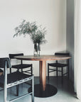A minimalist dining area features a SOBU Posta Dining Table - Walnut, paired with three black chairs and a glass vase with tall green branches, set against white walls and a concrete floor. Light filters in from the right. Custom sizes available.