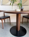 A round SOBU Posta Dining Table - Walnut with a black base stands between two wooden chairs with black seats. Stacked white dishes and a small potted plant rest on the table, with an abstract painting on the wall behind.