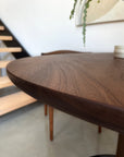 A close-up of the SOBU Posta Dining Table - Walnut, featuring smooth edges and a round solid walnut top, stands in a modern minimalistic room with a stairway, chair, plant, and wall art in the background; custom sizes offered.