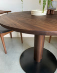 The SOBU Posta Dining Table - Walnut features a round solid walnut top and dark pedestal base, paired with a matching chair with a black seat. Two stacked white plates and a potted plant adorn the table. Custom sizes available.