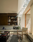 A sunlit modern living room featuring the Ethnicraft Eda Coffee Table, wooden shelves adorned with books and decor pieces, a trailing potted plant, a light wooden chair with European craftsmanship, glass doors opening outside, and sunlight streaming through a skylight.