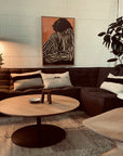 A cozy living room with a dark sectional, monochrome cushions, SOBU’s Disco Round Coffee Table in white oak, wooden chairs, a floor lamp, potted plant, and modern art on a white brick wall. Warm, soft lighting fills the space.