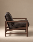 A side view of the Troubadour Lounge Chair | Leather by Wooden Stories, a modern armchair with dark brown leather cushions and a wooden frame, inspired by mid-century design, set against a plain beige background.