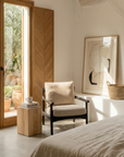 A cozy, sunlit bedroom features a cushioned chair, the SOBU Mars Terra Side Table, a woven basket, and abstract art. Large glass doors open to a patio with potted plants and a sculptural plant stand for added style.