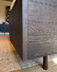 Close-up of a modern wooden sideboard with visible wood grain, next to the SOBU Woody Sofa (One Arm) in Espresso Ash and Fog. Set on a woven beige rug in a contemporary space with blurred artwork and furniture in the background.