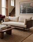 A Woody Sofa by SOBU, crafted in espresso ash and topped with natural kapok cushions, rests on a woven rug and faces a wooden coffee table adorned with books and a bowl. A vase of flowers and a large framed ocean photograph complete the cozy, sunlit living room—an embodiment of sustainable luxury furniture.