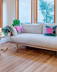 A modern living room features the SOBU Woody Sofa SAMPLE_(DZ) in light gray, a blue armchair, geometric pillows, plants, and solid ash wood furniture. Large windows add brightness; a pink mug and lamp are on small tables.