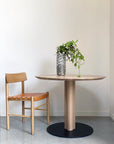 A SOBU Posta Dining Table - White Oak with a satin black steel base holds a tall vase of green and white foliage. Next to it, a light wood chair with a tan woven leather seat sits against a plain white wall.