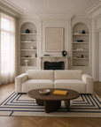 A modern living room featuring a cream Baker Sofa by Wooden Stories in fabric, paired with an abstract coffee table, striped rug, built-in shelves adorned with decorative objects, and a large framed blank artwork above a classic fireplace. Natural light streams through sheer curtains.