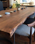 The Leviathan Live Edge Dining Table by SOBU, crafted from smoked acacia with curved metal legs, is set with small dishes and a potted plant. Modern wooden chairs with fabric seats surround it in a cozy, well-lit room.