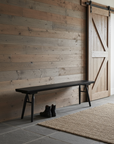 A minimalist room with a wooden plank wall, sliding barn door, SOBU’s Mars Live Edge Bench in Black Ash, black boots on the floor, and a woven rug atop gray stone tiles.