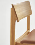 A close-up of Wooden Stories’ Poise Dining Chair in Light Oak with Leather highlights its modern Danish lines, curved backrest, and brown leather seat, set against a plain white background.