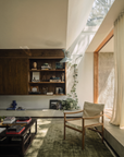 A cozy, sunlit living room featuring a green rug, wooden shelves filled with books and decor, the inviting Ethnicraft Safari Lounge Chair made of solid teak, abundant houseplants, and large windows adorned with sheer curtains that welcome in natural light.