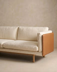 The Sonder Sofa | Fabric with Leather Back by Wooden Stories features modern cream-colored textured upholstery, wooden legs, and a tan leather panel on the armrest from the Danish collection, set against a neutral beige wall and floor.