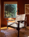 A SOBU Viking Lounge Chair - Black Ash sits on a cowhide rug in a sunlit room with wood-paneled walls, a large window framed by bamboo, and abstract wall art.