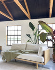 A minimalist living room features the SOBU Woody Sofa (One Arm) in white ash with natural kapok cushions, accented by a green throw. Large leafy plants sit beside the sofa, sunlight pours in through grid windows, and exposed wood beams highlight the ceiling.