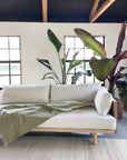The SOBU Woody Sofa (One Arm) in White Ash with Natural Kapok hypoallergenic cushions and an olive green throw blanket sits before a white wall with large windows, tall leafy plants behind, and wooden ceiling beams overhead.