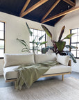 A modern living room featuring the SOBU Woody Sofa (One Arm) in White Ash with Natural Kapok Cushions, paired with a green throw blanket, leafy plants behind the sofa, and natural light streaming through black-framed windows below exposed beams.