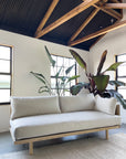A modern, minimalist living room features the SOBU Woody Sofa (One Arm) in white ash with natural kapok cushions, set against large indoor plants, exposed wooden beams, black ceiling panels, white walls, and natural light from two windows.